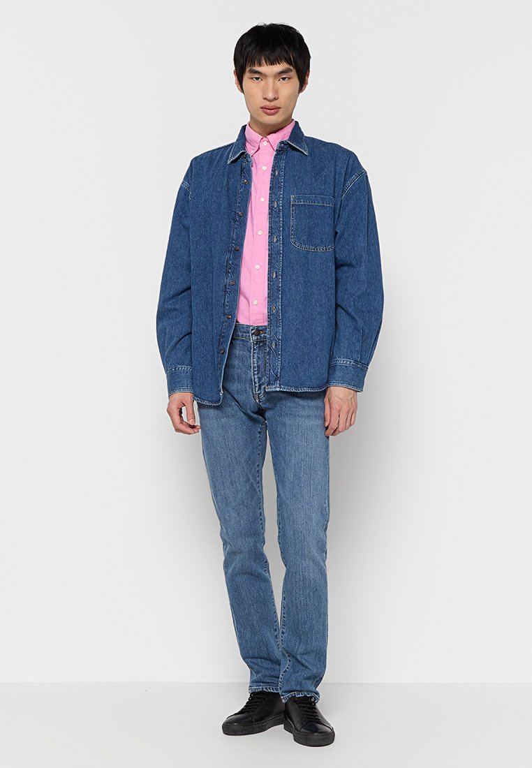 Chaqueta de mezclilla en azul oscuro sobre una camisa de botones rosa claro, combinada con jeans de azul medio y zapatos negros, con detalle en el bolsillo delantero.