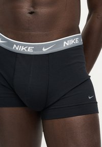 Άνω μέρος σώματος σκοτεινής επιδερμίδας άνδρα που φοράει μαύρο boxer Nike με γκρι ζώνη μέσης με λογότυπο Nike και swoosh, στέκεται σε λευκό φόντο.