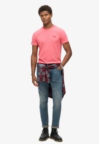 Rød t-shirt, blå denim jeans, ternet skjorte bundet om taljen, sorte snørebåndsstøvler. Ur på håndleddet, stående i en neutral stilling.