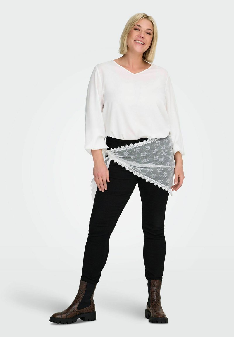 Femme souriante aux cheveux blonds porte un chemisier blanc, un pantalon noir, un cache-cœur en dentelle noué à la taille et des bottines marron à motifs, debout sur fond blanc.