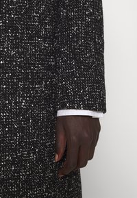 Svart och vit texturerad tygkappa, med ett vävt mönster och en vit skjortärm synlig vid ärmen.