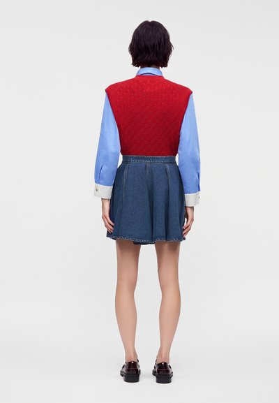 Femme debout de dos, portant un pull sans manches rouge sur une chemise bleu clair à manches longues, une jupe en jean bleue et des chaussures foncées.
