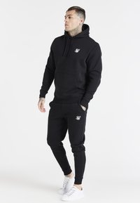 Ensemble en mélange de coton comprenant un sweat à capuche noir et un jogging, avec une poche avant, des poignets côtelés et un petit logo blanc. Associé à des baskets blanches.