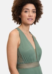 Zielona sukienka z głębokim dekoltem w kształcie litery V, koronkowymi panelami bocznymi oraz dekoracyjnym wykończeniem w talii. Miękka tkanina o gładkiej teksturze.