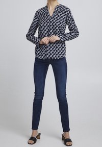 Navy blouse with white abstract pattern, V-neck, long sleeves, and fitted shape. Paired with dark skinny jeans and black block-heeled sandals.