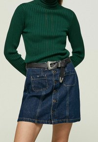 Un pull à col roulé en maille côtelée vert foncé associé à une jupe en denim bleu clair dotée de deux poches avant et d'une ceinture noire avec une boucle en argent.