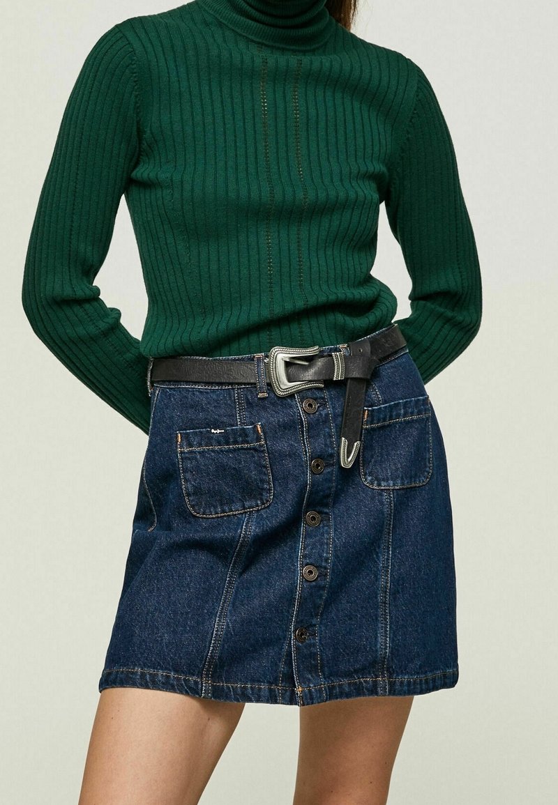 Un pull à col roulé en maille côtelée vert foncé associé à une jupe en denim bleu clair dotée de deux poches avant et d'une ceinture noire avec une boucle en argent.