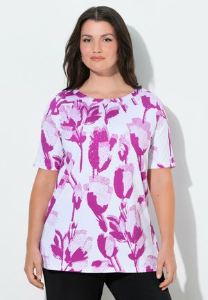 Mujer con cabello largo y oscuro que lleva una camiseta blanca de manga corta con audaces estampados florales morados, de pie contra un fondo liso y claro.