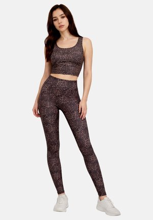 Woman in leopard print workout crop top and leggings with white sneakers standing against plain background.