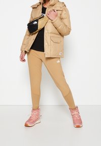 Osoba v béžové péřové bundě, černém tričku, béžových legínách, růžových zimních botách a černobéžové křížové tašce stojí na bílém pozadí.