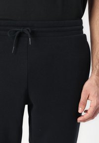 Pantaloni de trening negri, confecționați dintr-un material moale, cu talie ajustabilă prin șnur cu capete metalice și buzunare laterale. Design minimalist, fără modele vizibile.