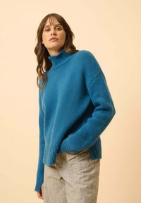 Niebieski sweter z dzianiny z wysokim kołnierzem, w luźnym kroju i ściśniętymi mankietami. W połączeniu z jasnymi, strukturalnymi spodniami.