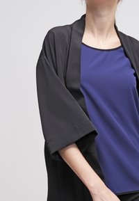 Černý kimono kabát s širokými rukávy, nošený přes modrý top. Kabát má hladkou strukturu a uvolněný střih, který zdůrazňuje moderní design.