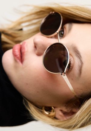 Gros plan d'une personne aux cheveux blonds portant des lunettes de soleil rondes et des boucles d'oreilles en or en forme d'anneau, regardant légèrement vers le haut et sur le côté.