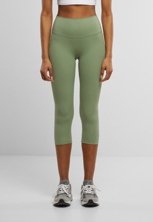 Leggings visokog struka u maslinastoj boji, izrađene od rastezljivog materijala, sa glatkom teksturom i skraćenom dužinom, u kombinaciji sa sivim sportskim cipelama.