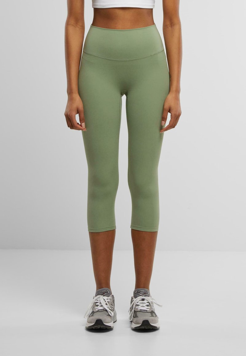 Hoge taille, olijf groene leggings van rekbaar materiaal, met een gladde textuur en een korte lengte, gecombineerd met grijze sportschoenen.