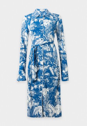 Woman wearing a blue and white long-sleeve belted midi dress with abstract nature patterns and yellow high-heeled shoes standing against white background.