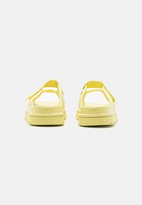 Sandales jaunes avec une semelle épaisse, un dos ouvert et des brides ajustables. Le matériau semble lisse avec une finition mate et des accents texturés.