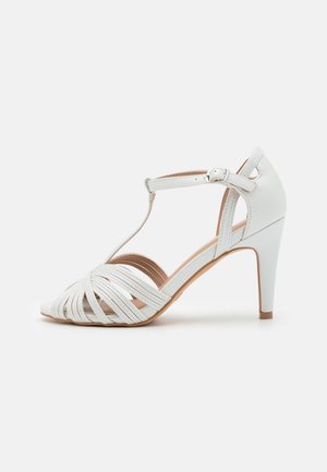 Sandale blanche à talons hauts avec bride en T, fermeture à boucle et lanières avant en cuir tressé, vue de profil sur un fond uni.