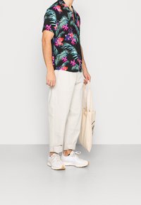 Chemise noire à manches courtes avec des imprimés floraux colorés et des feuilles de palmier, associée à un pantalon ample beige clair et des baskets blanches. Sac fourre-tout en coton inclus.
