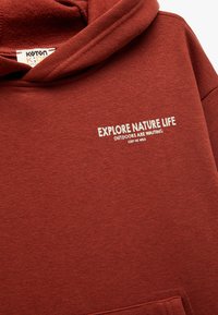 Hoodie in gebrandoranje, gemaakt van zachte stof. Voorzien van witte tekst: "ONTDEK DE NATUURLIJKE LEVEN," "BUITEN WACHTEN," en "HOU MIJ WILD."