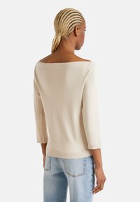 Světle béžový svetr bez ramínek s tříčtvrtečními rukávy, žebrovanými manžetami a hladkou texturou, v kombinaci se světle modrými džínami.