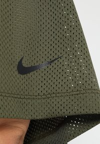Zielona siatkowa koszulka sportowa z czarnym logotypem Nike. Posiada małe otwory zapewniające wentylację oraz zaokrąglony dół. Tekstura materiału wydaje się lekka.