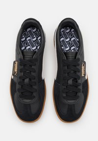 Baskets en daim et cuir noir avec une semelle en caoutchouc marron, dotées de lacets noirs et d'une bande blanche sur le côté. L'intérieur comporte une semelle intérieure à motif.