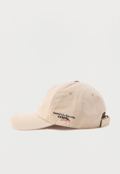 Casquette de baseball beige avec une visière courbée et une sangle réglable, ornée d'un texte brodé noir et rouge sur le panneau latéral.