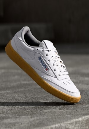 Valkoinen nahkainen tennari, jossa on keltainen, kuvioitu kumipohja. Sivulla sininen Reebok-logo ja kärjessä rei'ityksiä.