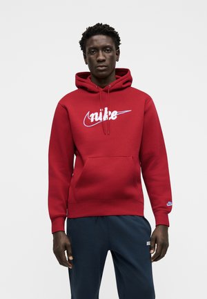 Młody mężczyzna stoi, ubrany w czerwoną bluzę z kapturem Nike z białym logo i granatowe spodnie dresowe Nike na jednolitym białym tle.