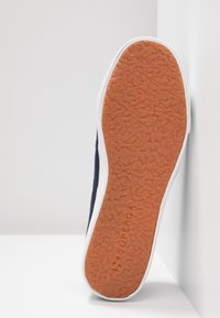 Podešev navy modrého tenisu s texturovanou oranžovou gumovou podrážkou a bílou mezipodešví, umístěná svisle na bílém pozadí.