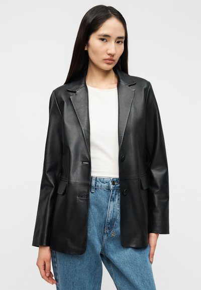 Μαύρο δερμάτινο σακάκι με λοξές πέτρες γιακά, κλείσιμο με ένα κουμπί και δύο εμπρόσθιες τσέπες, συνδυασμένο με ανοιχτόχρωμο τζιν και λευκό τοπ.