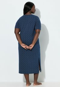 Mujer de pie descalza con las manos entrelazadas detrás de la espalda, vistiendo un vestido midi azul marino con patrones circulares y una abertura lateral.