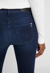 Jeans ajustados de color azul oscuro con una textura suave, que presentan dos bolsillos traseros, uno con un diseño bordado y botones decorativos.