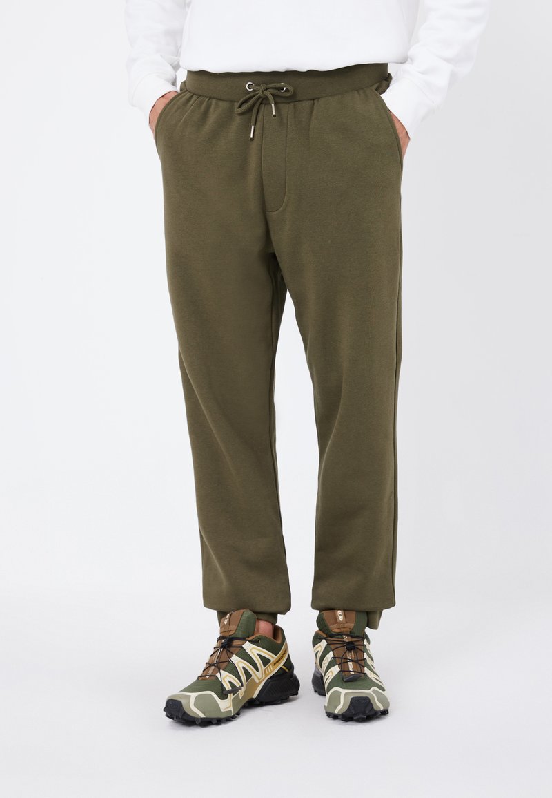 Olivengrønne joggers med elastisk talje, snorelukning, sidelommer og ribkant ved anklerne, parret med strukturerede sneakers.