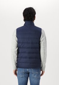 Gilet matelassé bleu marine avec un col haut, comportant des sections rembourrées. Porté par-dessus un pull gris clair, avec une texture lisse et isolante.