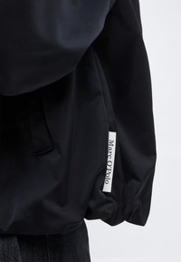 Črni rokav jopiča z belo tkanino etiketo z napisom "Marc O'Polo" na svetlem ozadju.