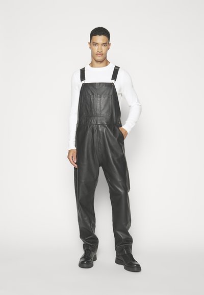 Zwarte leren overall met verstelbare banden, een voorzak en een losse pasvorm; gecombineerd met een lange mouw witte top en zwarte schoenen.