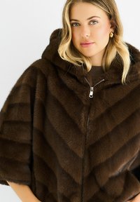 Woman with blonde hair wearing gold hoop earrings and a dark brown fur coat with a hood and visible silver zipper.