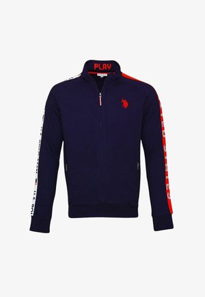 Mann trägt eine marineblaue Jacke mit Reißverschluss, roten Streifen und Logo an Ärmeln und Brust, blaue Jeans und schwarze und weiße Turnschuhe.