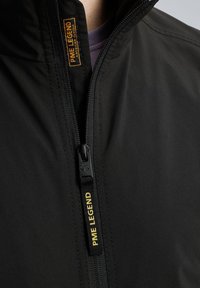 Schwarze Jacke mit einem vertikalen Reißverschluss, strukturiertem Stoff und einem Etikett mit "PME LEGEND" in Gelb. Nahaufnahme konzentriert sich auf den Reißverschluss und den Kragen.