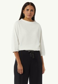 Witte geprofileerde trui met een hoge, ronde hals en driekwartmouwen, gecombineerd met zwarte high-waisted broek met een strikceintuur.