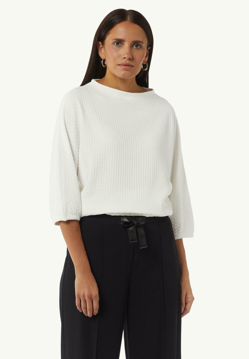 Witte geprofileerde trui met een hoge, ronde hals en driekwartmouwen, gecombineerd met zwarte high-waisted broek met een strikceintuur.