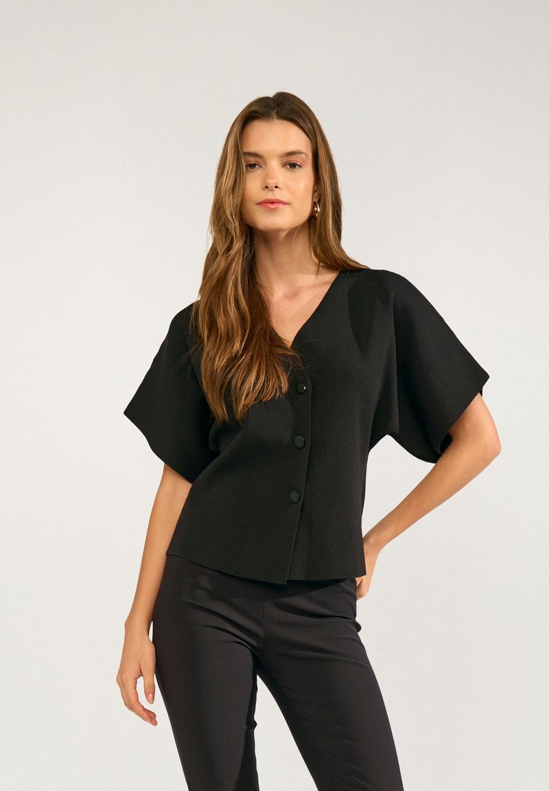 Femme aux longs cheveux bruns portant un haut noir à col en V boutonné avec de larges manches courtes et un pantalon noir moulant, debout avec une main sur la hanche.