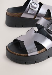 Strieborné metalické sandále na platforme s čiernymi remienkami a korkovou stielkou, vybavené nastaviteľnou prackou a textúrovanou gumovou podrážkou.