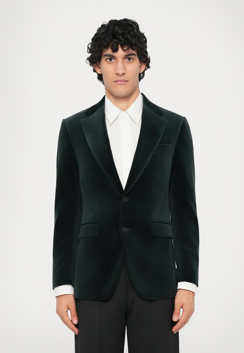 Zelený sametový blazer s klopovými límci, dvěma předními kapsami a jedním knoflíkem, na sobě s bílou košilí s límečkem.