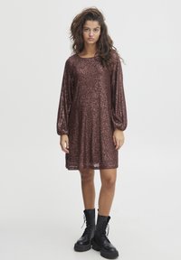 Robe en sequins bronze avec de longues manches bouffantes, un col rond et une coupe ample ; associée à des bottes de combat noires à lacets. Surface texturée et réfléchissante.