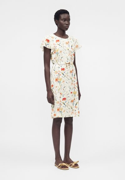 Γυναίκα που φοράει λευκό φόρεμα μέχρι το γόνατο με πορτοκαλί και κίτρινα λουλουδένια σχέδια, κοντά φουσκωτά μανίκια, λεπτή χρυσή ζώνη και μπεζ σανδάλια με φιόγκους.