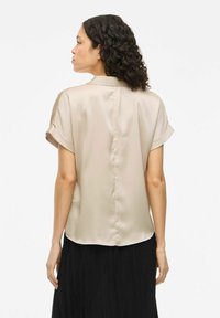 Top in satin leggero beige chiaro a maniche corte con un leggero luccichio, caratterizzato da una vestibilità dritta e maniche arrotolate, abbinato a una gonna nera plissettata.